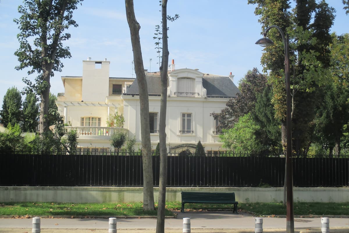Maison blanche à toit d'ardoise derrière une longue grille noire et des arbres, à l'angle d'un boulevard planté.