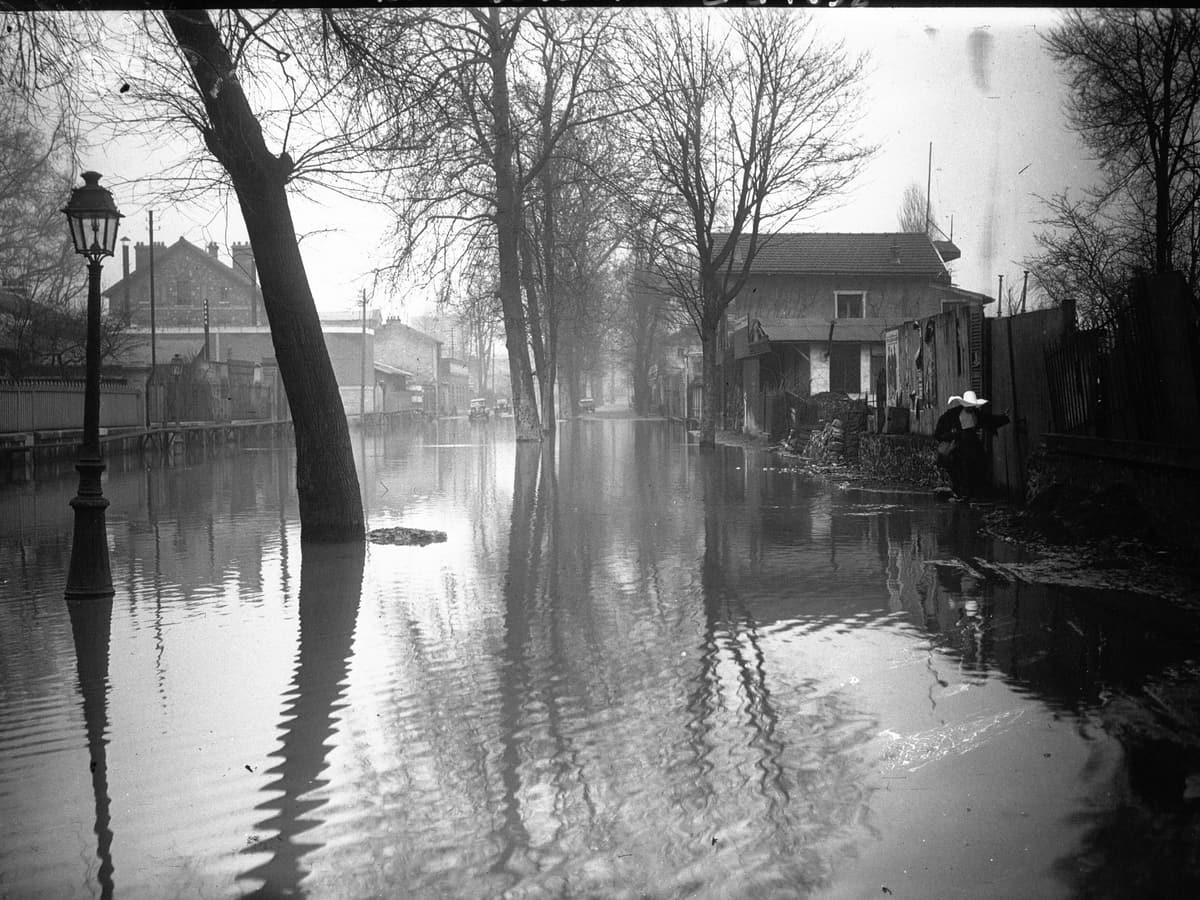 Photographie de 1926 : une rue inondée, l'eau monte au pied des réverbères et des arbres, des maisons basses au fond.