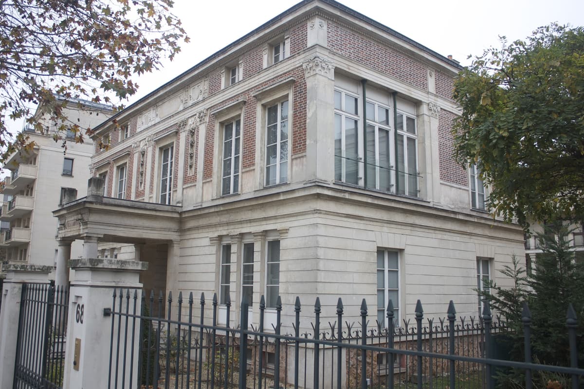 Hôtel particulier en brique et pierre derrière une grille, avec un porche à colonnes, sous les feuillages d'automne.