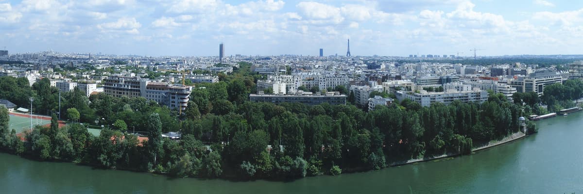 Panorama de Neuilly-sur-Seine depuis l'autre rive : la Seine au premier plan, l'île de la Jatte boisée, puis la ville et Paris au loin.