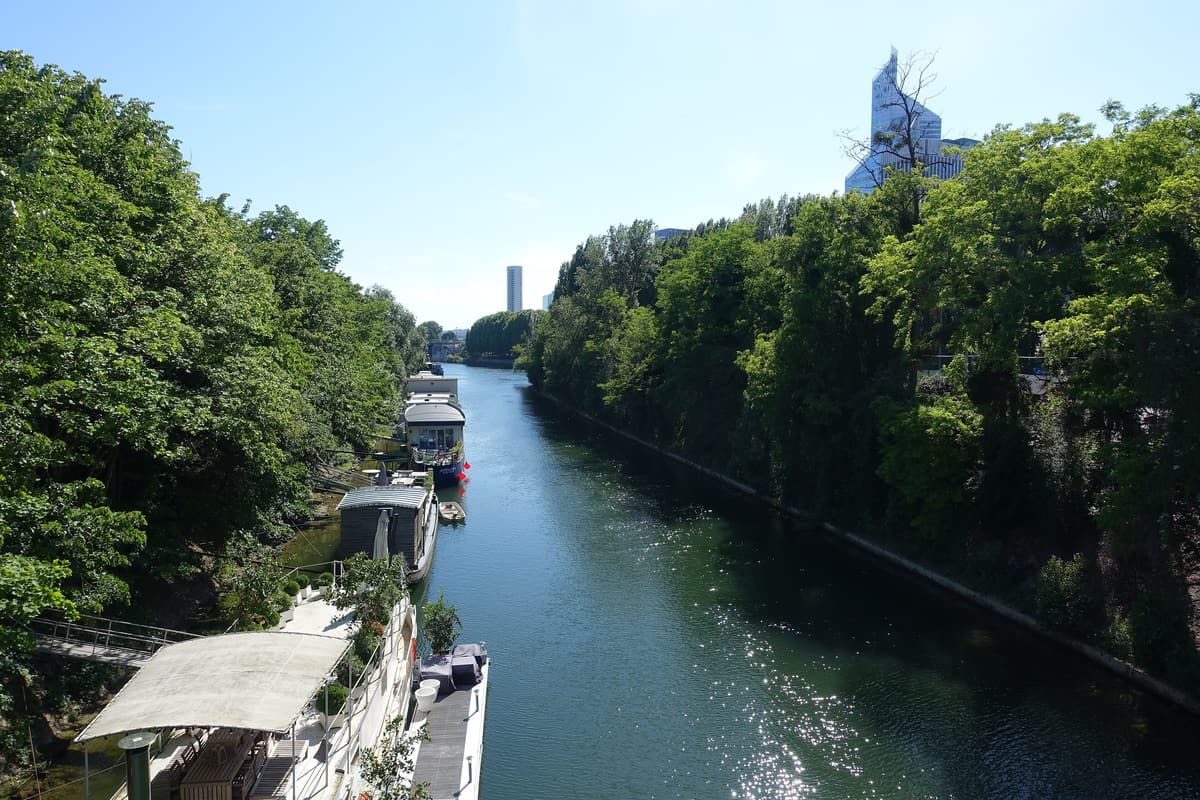 Un bras de la Seine entre deux rives boisées, des péniches amarrées sur la gauche.